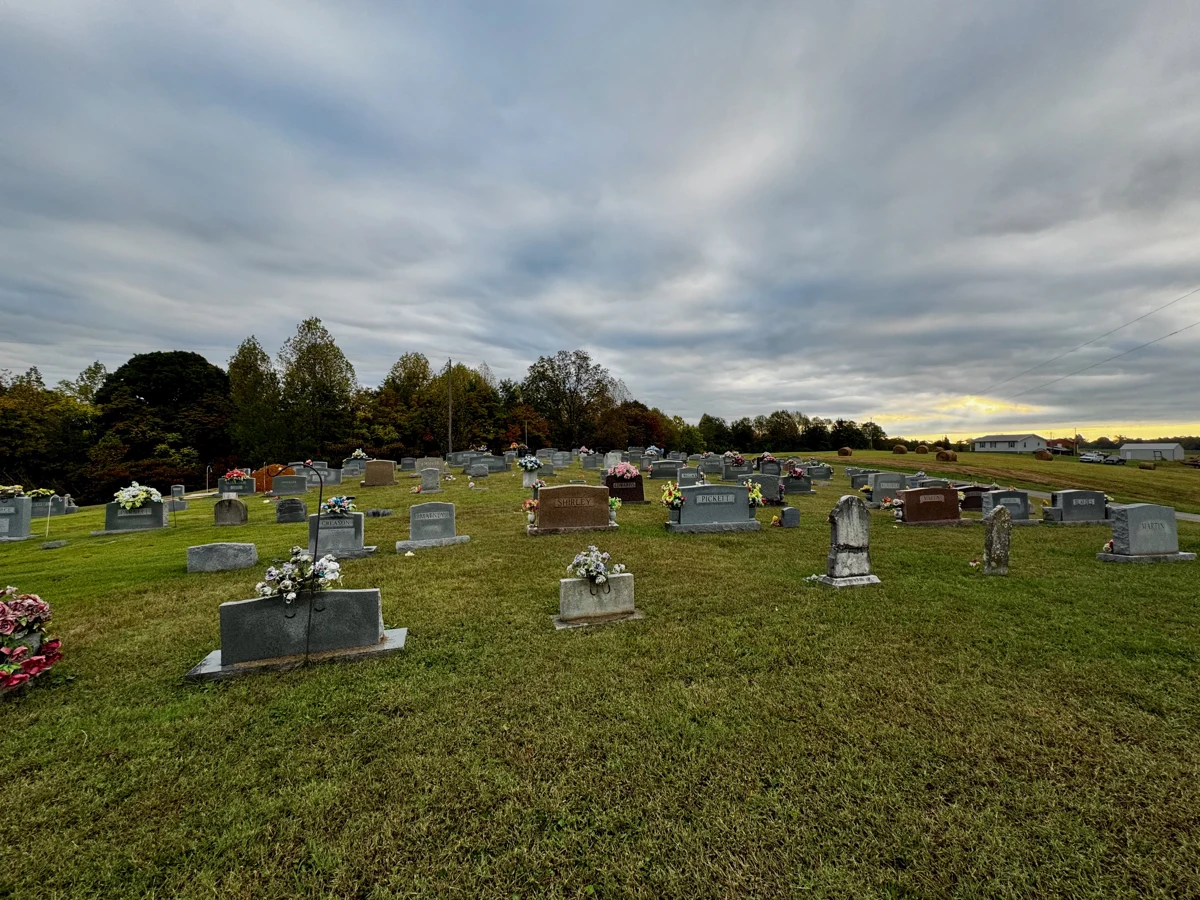 The mown cemetery grounds and flower-decorated monuments, with fields and hay bales beyond.